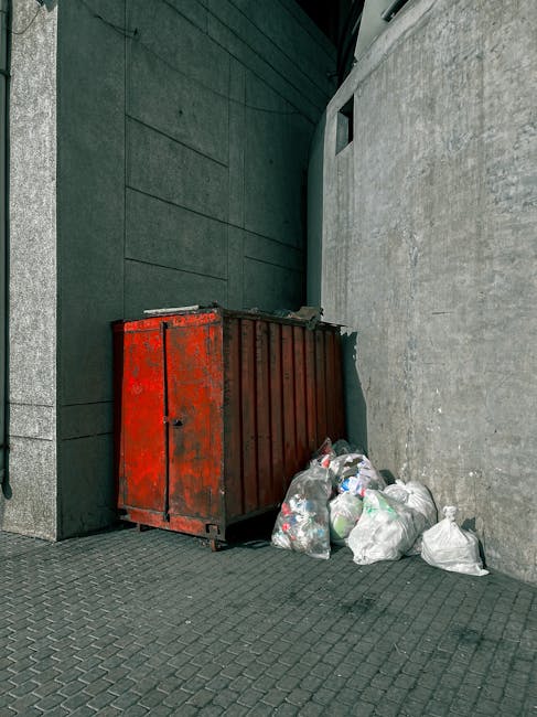 A row of large black wheelie bins lined up along a residential pavement beside red-brick terraced houses, with some bins showing white spray-painted numbers. A person dressed in a brown coat is seen p