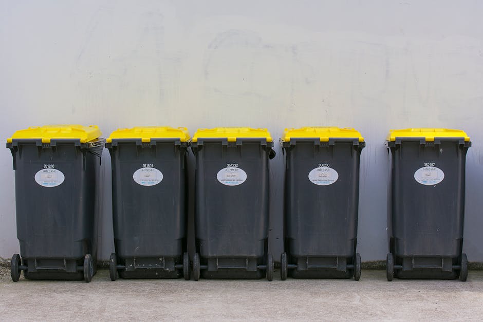 A row of five large black wheeled rubbish bins with yellow lids, aligned against a plain light gray wall outdoors. Each bin has a white oval label and a unique identification number printed on the fro